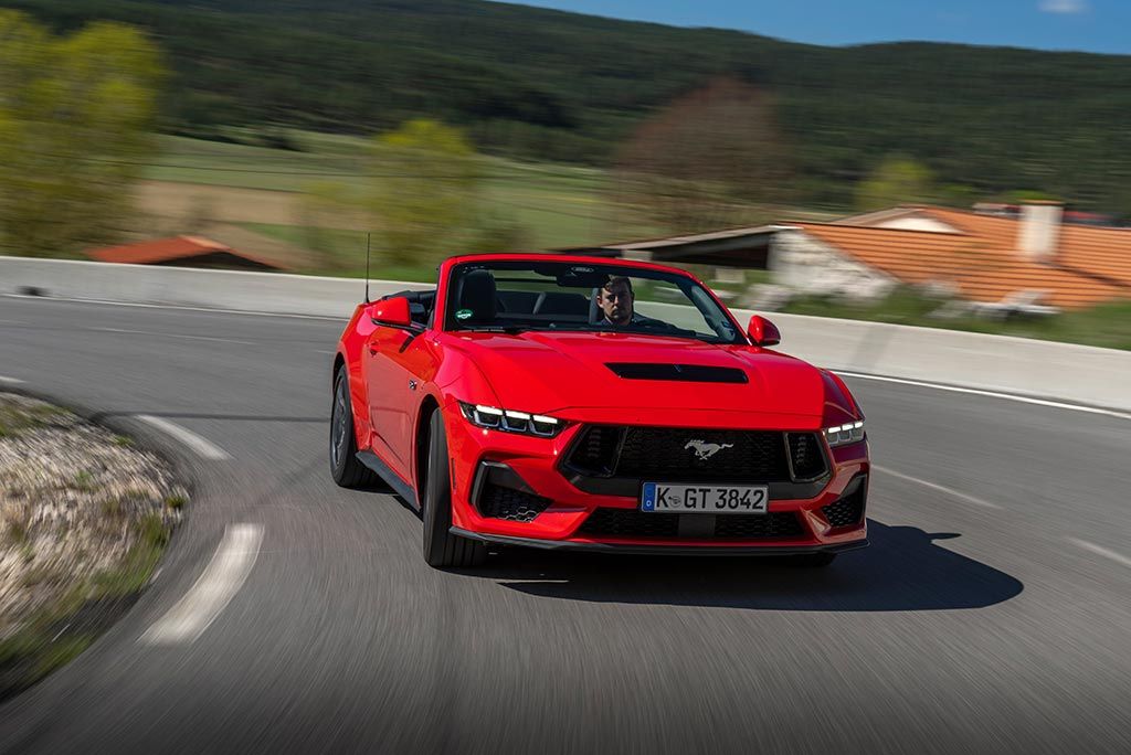 Roter Ford Mustang Cabriolet fährt auf kurviger Landstraße mit bewaldeten Hügeln im Hintergrund.
